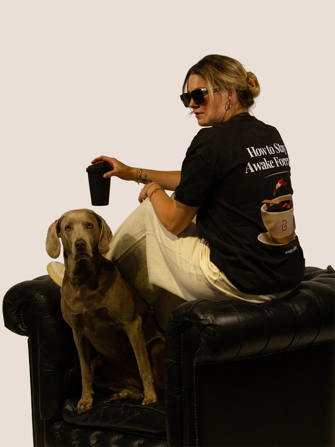 Frau im schwarzen Kaffee-Heavy-Tee von Bengel & Göre auf Ledersessel mit Weimaraner-Hund, Rückenprint.
