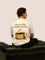 Mann im Off-White „How To Stay Awake Forever“ Heavy Tee auf schwarzem Ledersofa, Kaffeetassen-Rückenprint.