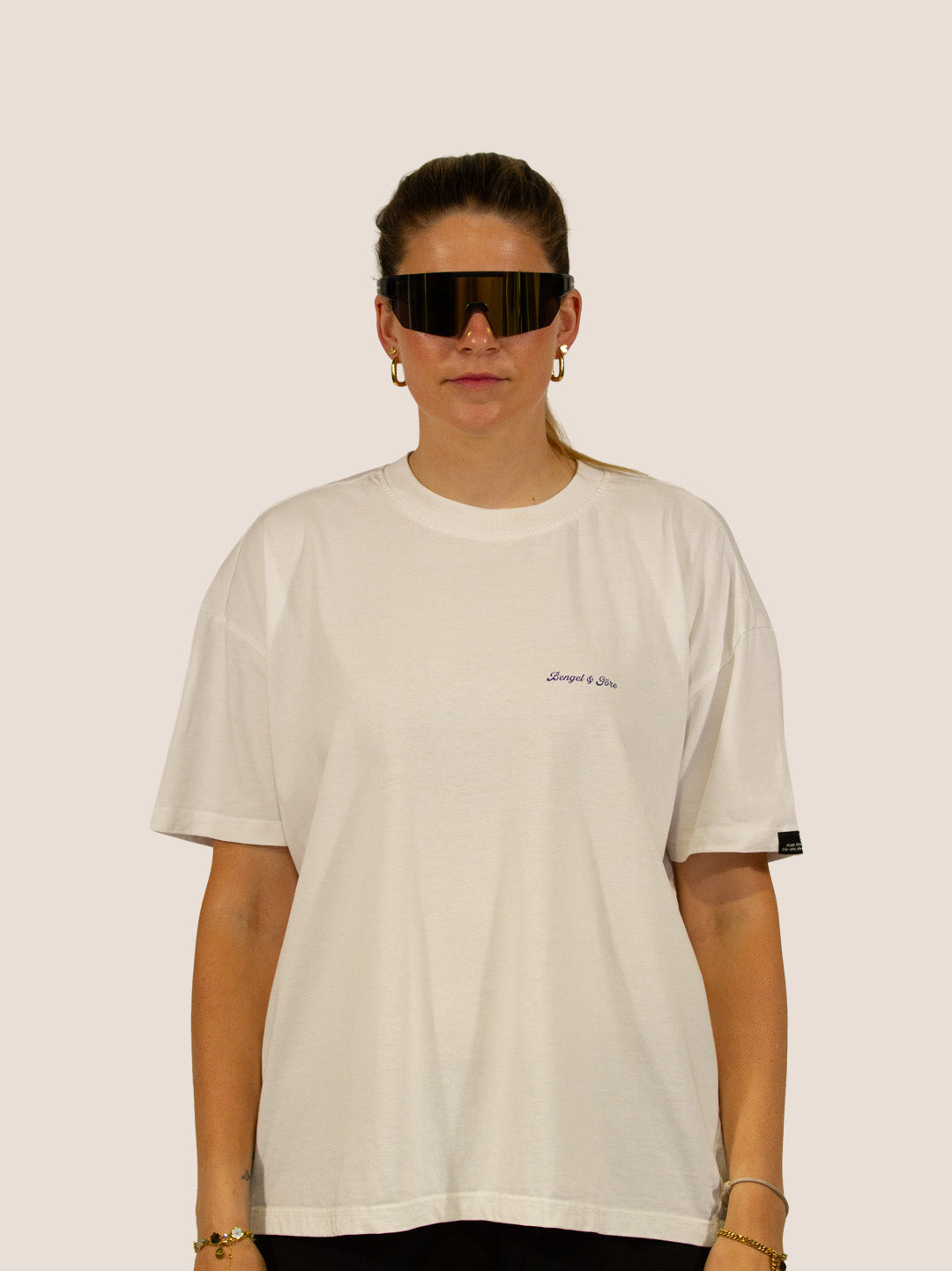 Frau im Off-White Heavy Tee von Bengel & Göre mit Brust-Schriftzug und Rennrad-Sonnenbrille, Vorderseite.