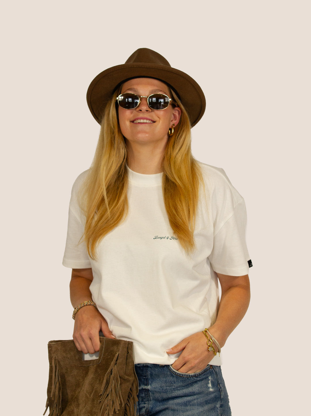 Lächelnde Frau im Off-White Heavy Tee von Bengel & Göre mit Filzhut und Wildleder-Fransentasche, Western-Look.