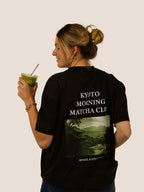 Frau im schwarzen „Kyoto Morning Matcha Club“ Heavy Tee von Bengel & Göre mit Kyoto-Landschaftsprint, hält Matcha.