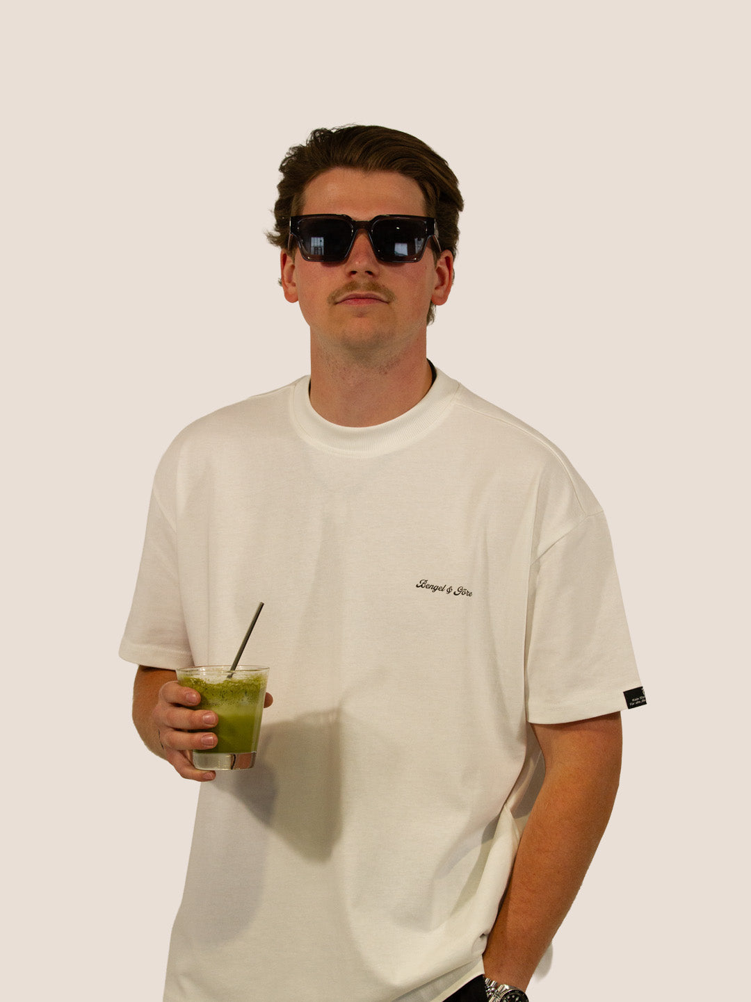 Mann im Off-White Bengel & Göre Heavy Tee mit Sonnenbrille und Matcha-Getränk, Brust-Schriftzug, Vorderseite.