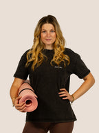 Frau im schwarzen Bengel & Göre Heavy Tee mit Brust-Schriftzug und rosa Pilatesmatte, Vorderseite.