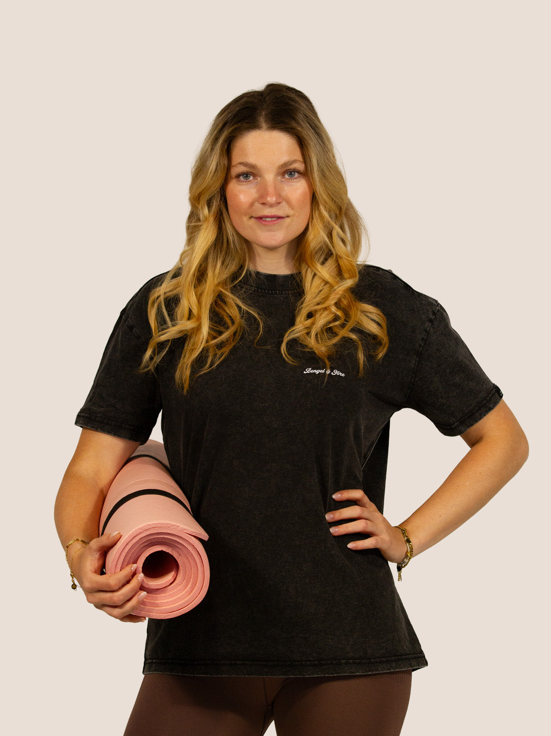 Frau im schwarzen Bengel & Göre Heavy Tee mit Brust-Schriftzug und rosa Pilatesmatte, Vorderseite.