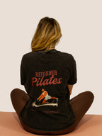 Frau im schwarzen „Reformer Pilates“ Heavy Tee mit Reformer-Print, sitzend auf Matte, Rückansicht.