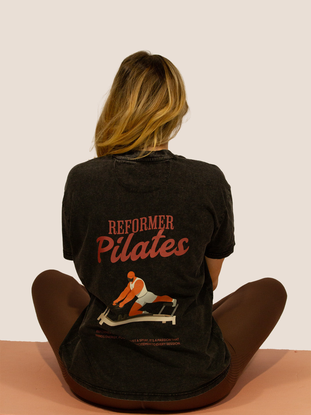 Frau im schwarzen „Reformer Pilates“ Heavy Tee mit Reformer-Print, sitzend auf Matte, Rückansicht.