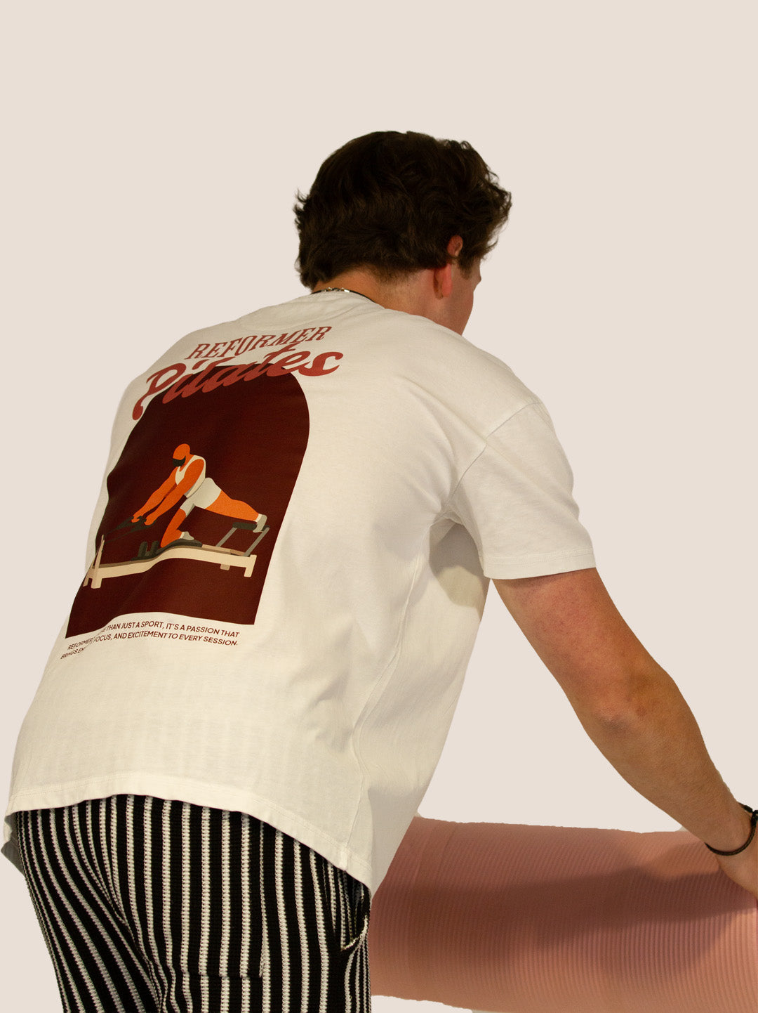 Mann im Off-White „Reformer Pilates“ Heavy Tee von Bengel & Göre, Rückenprint, mit rosa Pilatesmatte.